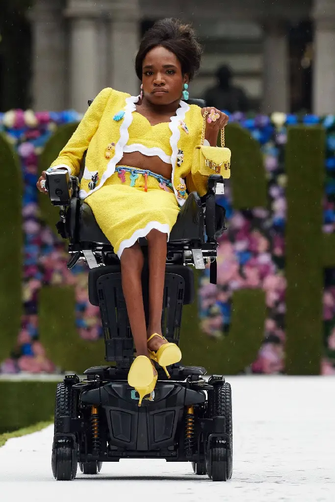 Woman in a wheelchair on a runway; she is wearing a canary yellow suit with a tube top, midi pencil skirt, and long sleeve jacket, each with a white scalloped edge. Her jacket is decorated with large stylized animal buttons, and her belt is made of colorful keys. Her open toed heels and small purse match her yellow suit. The purse is embellished with large gold charms spelling &#39;moschino&#39; dangling from the purse flap. Her short black hair is parted in the center in front and teased up in the back.