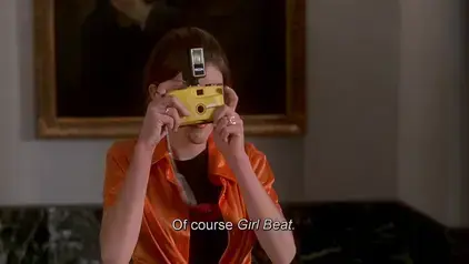 Screenshot from the movie Kids in the Hall: Brain Candy (1996), a young journalist taking a photograph during a small press conference, subtitle has part of the line thanking the few press attendees: &quot;Of course *Girl Beat.*&quot;