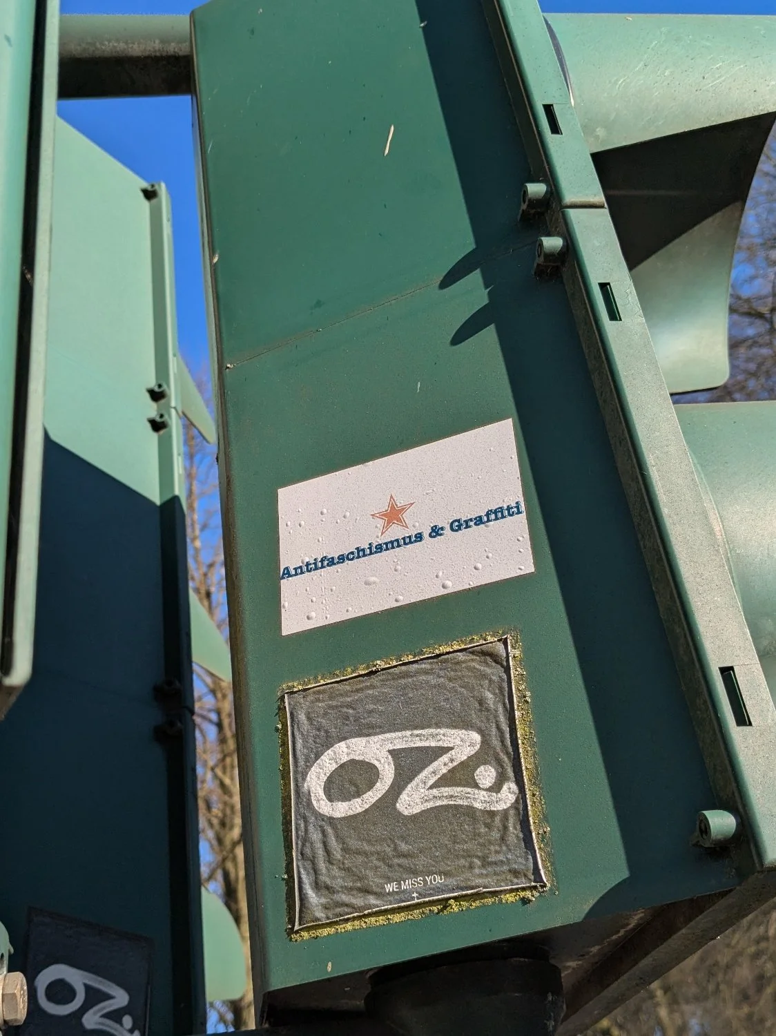 An einer Ampel kleben seitlich zwei Sticker. Auf dem ersten steht "Antifaschismus & Graffiti", auf dem zweiten "OZ we miss you"