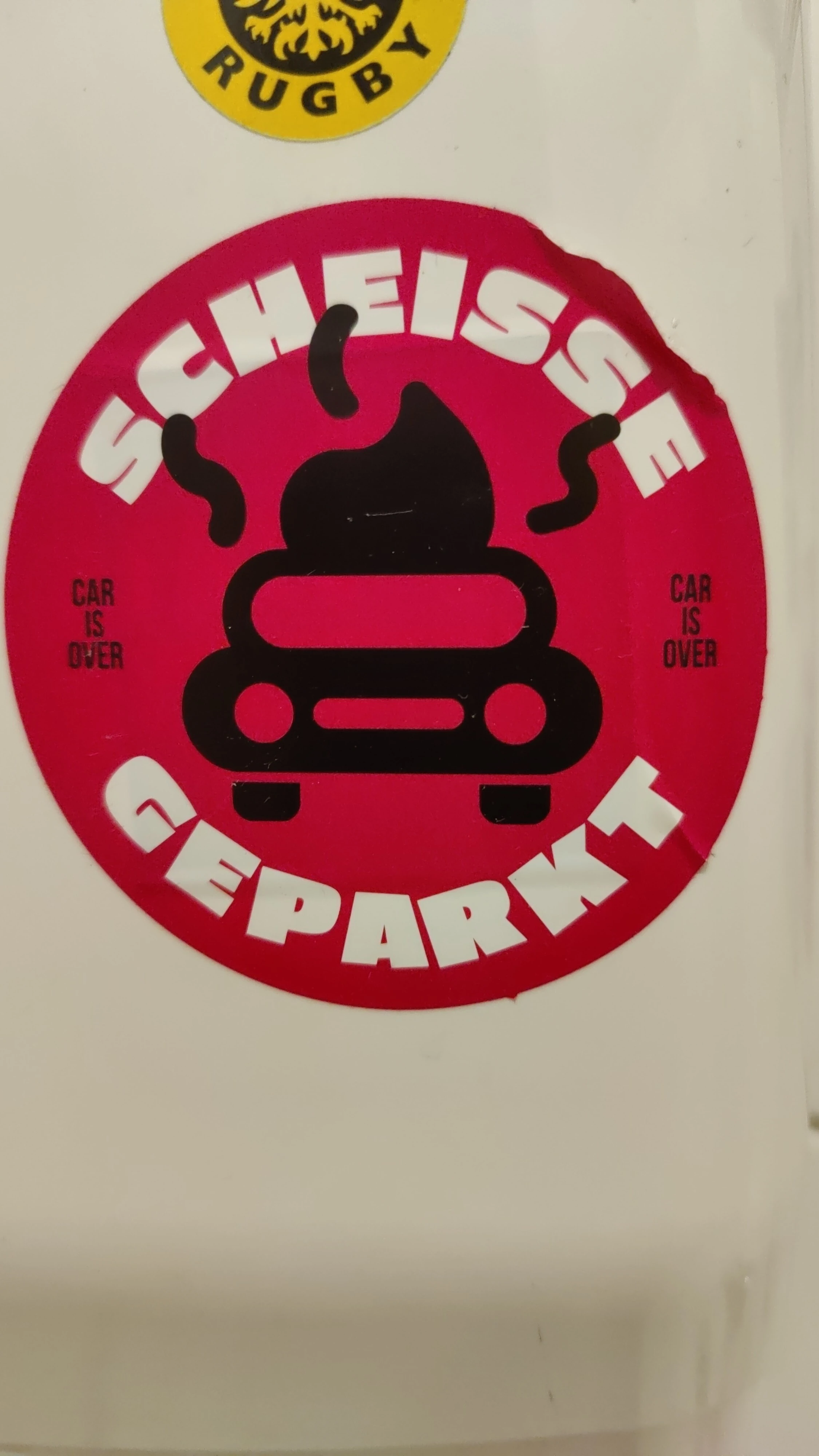 A round magenta sticker on a paper towel dispenser, depicting a car as a smelly poop with a legend in white saying scheiße geparkt (shit parked/ poorly parked) and "car is over" small on the sides