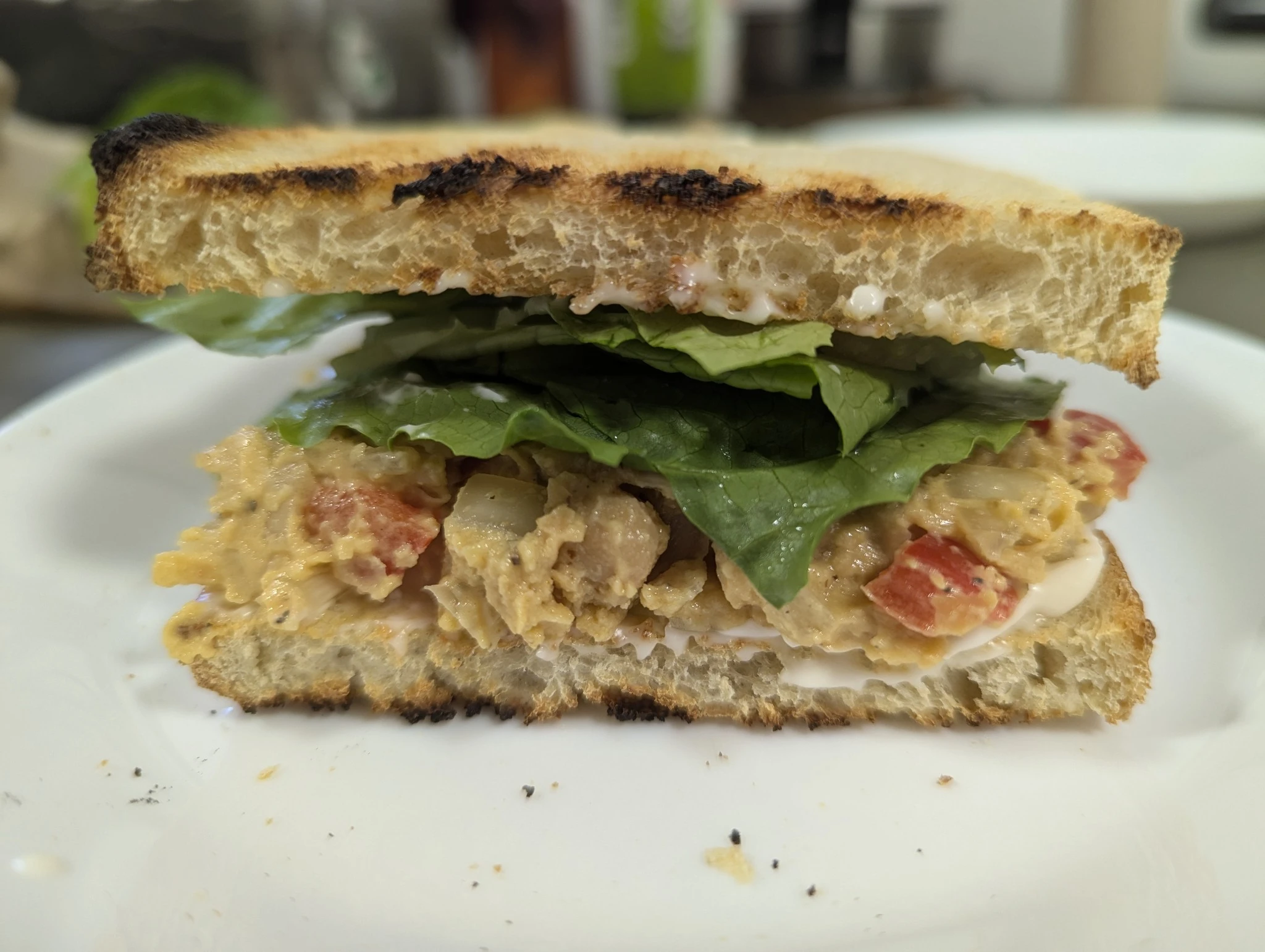 image contains a sandwich with toasted bread, lettuce, and a chickpea salad filling on a white plate and blurred background.