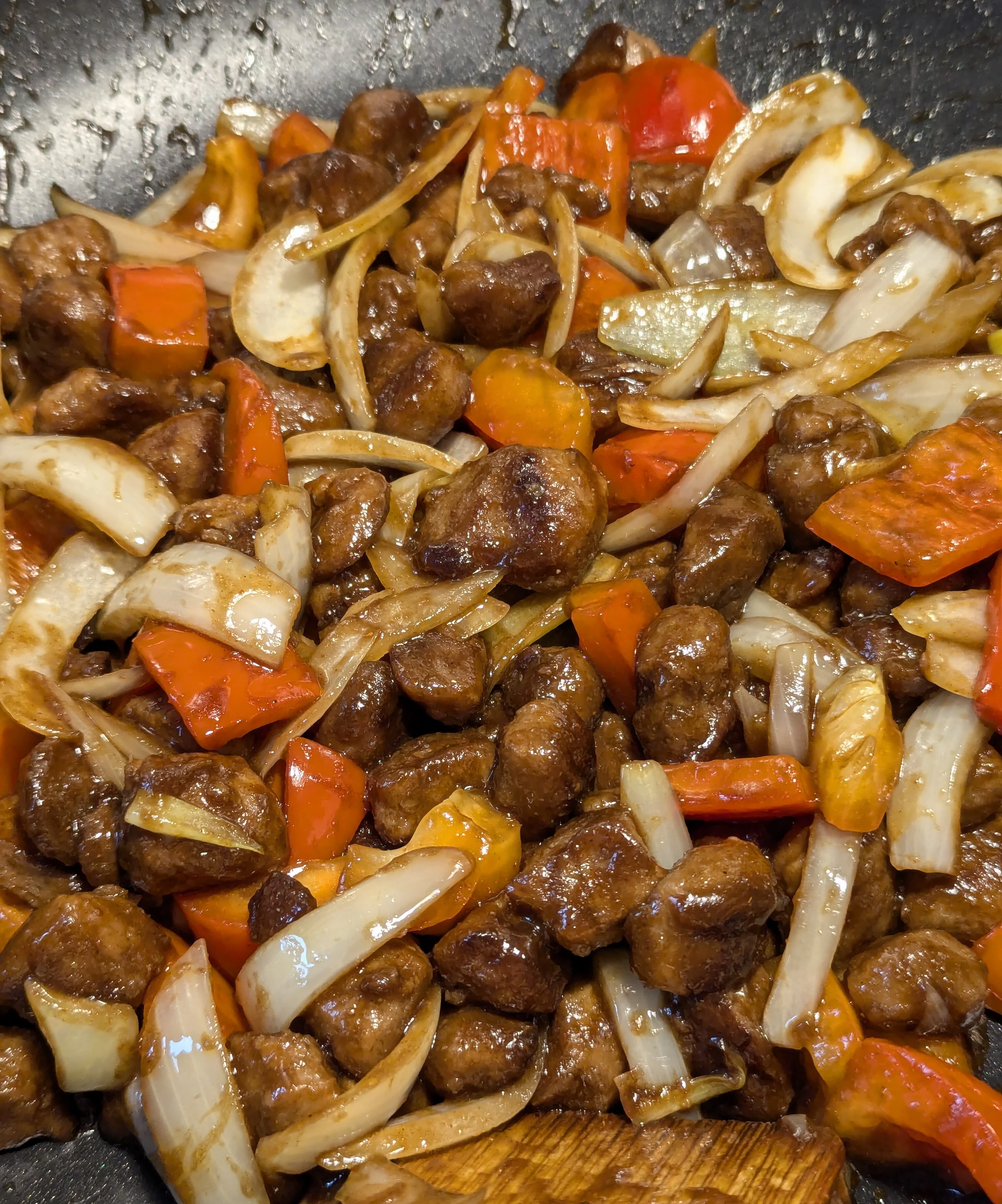 image is a close up of soya chunks, onion slices, and chopped red pepper that appears stir fried, covered in sauce, and is sitting in a wok.