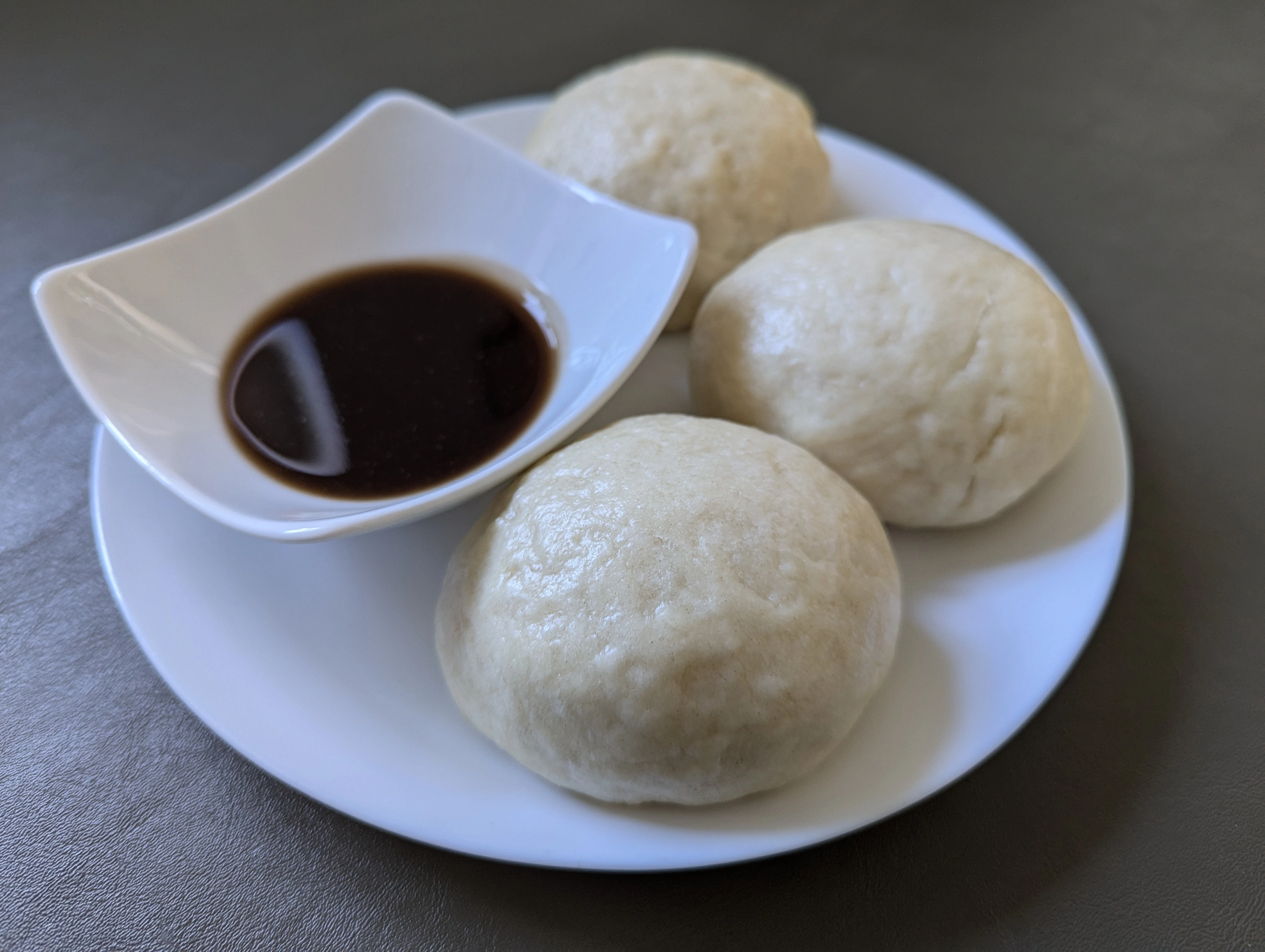 three siopao (steamed buns) arranged on a white plate with a small sauce bowl filled with a dark dipping sauce.