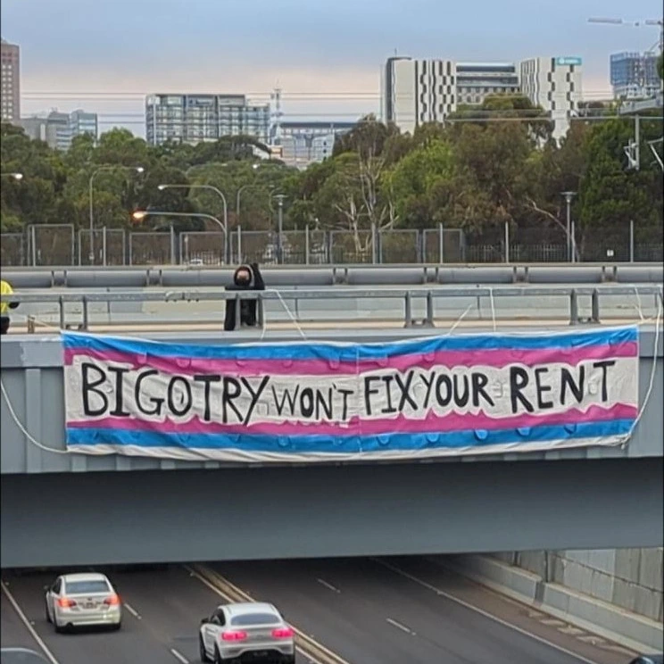 photo of a banner hung over a highway, a person dressed in black raises their fist in solidarity