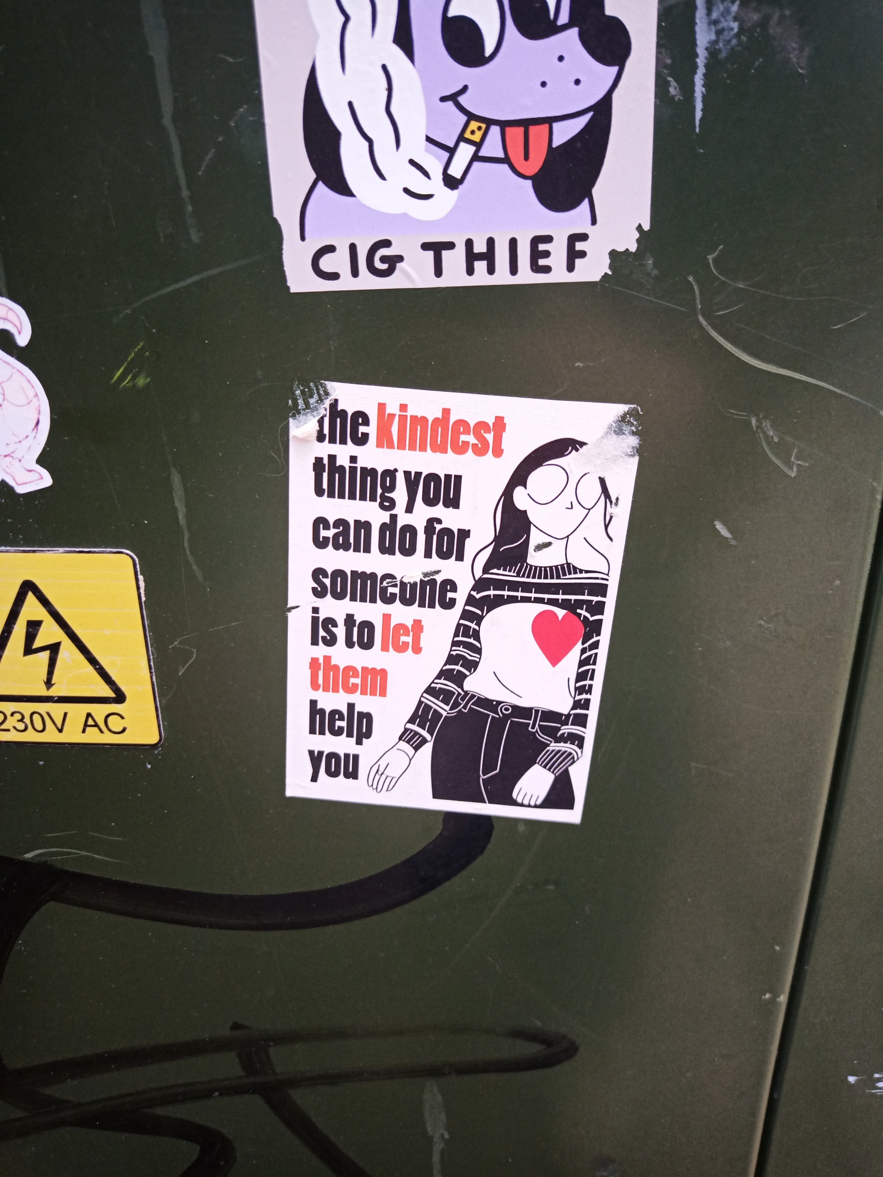 A sticker on a green utility box that has an illustration of a person with a long sleeve jumper rolled up above their chest and a red heart overlayed over one side of their chest. it has text next to it reading "the kindest thing you can do for someone is to let them help you"