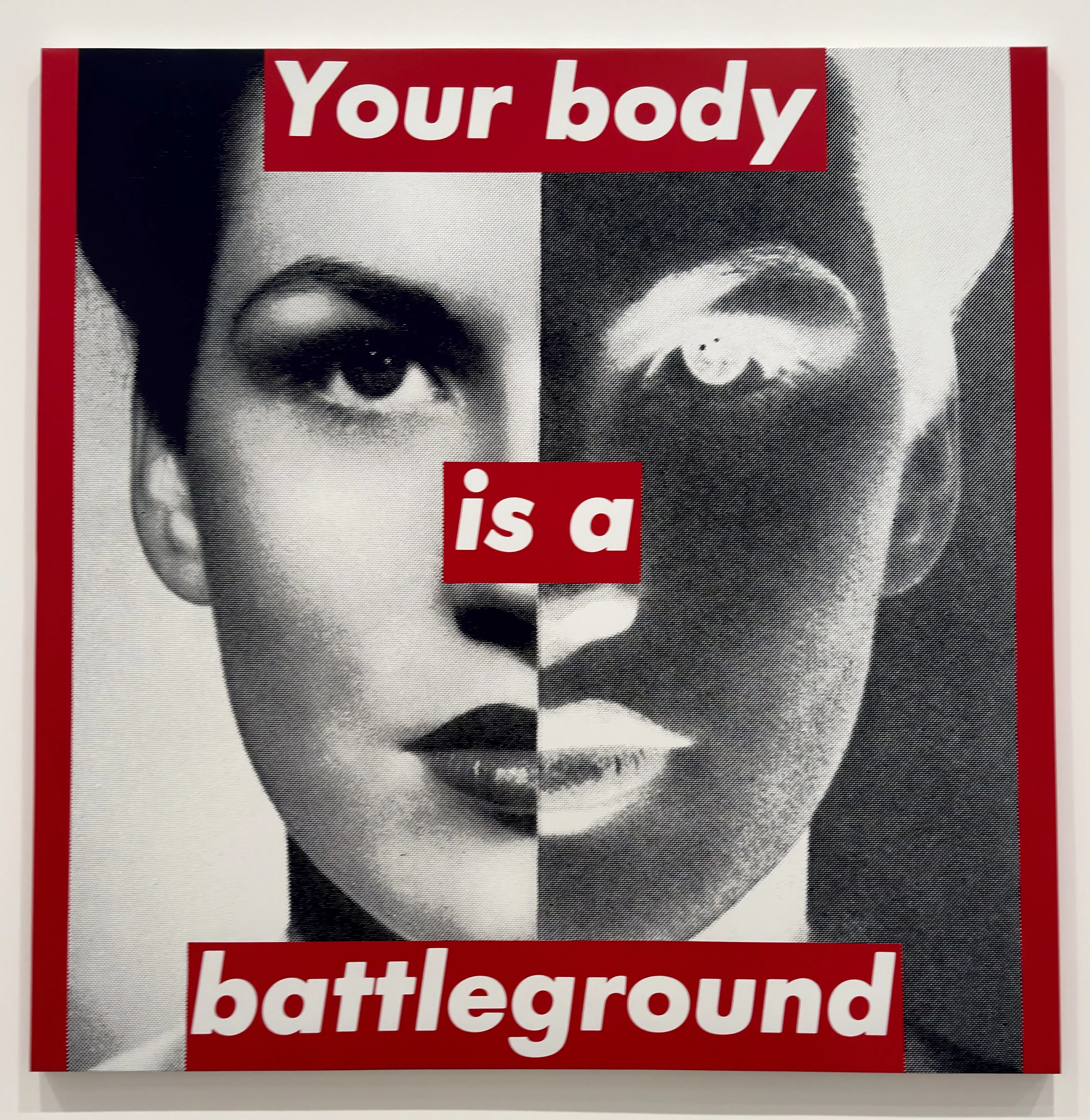 A photograph of a woman that is split down the middle, with half a normal black and white photograph, and the other half inverted. The photo is bordered on both sides by bright red, and white text on bright red says “Your body is a battleground”