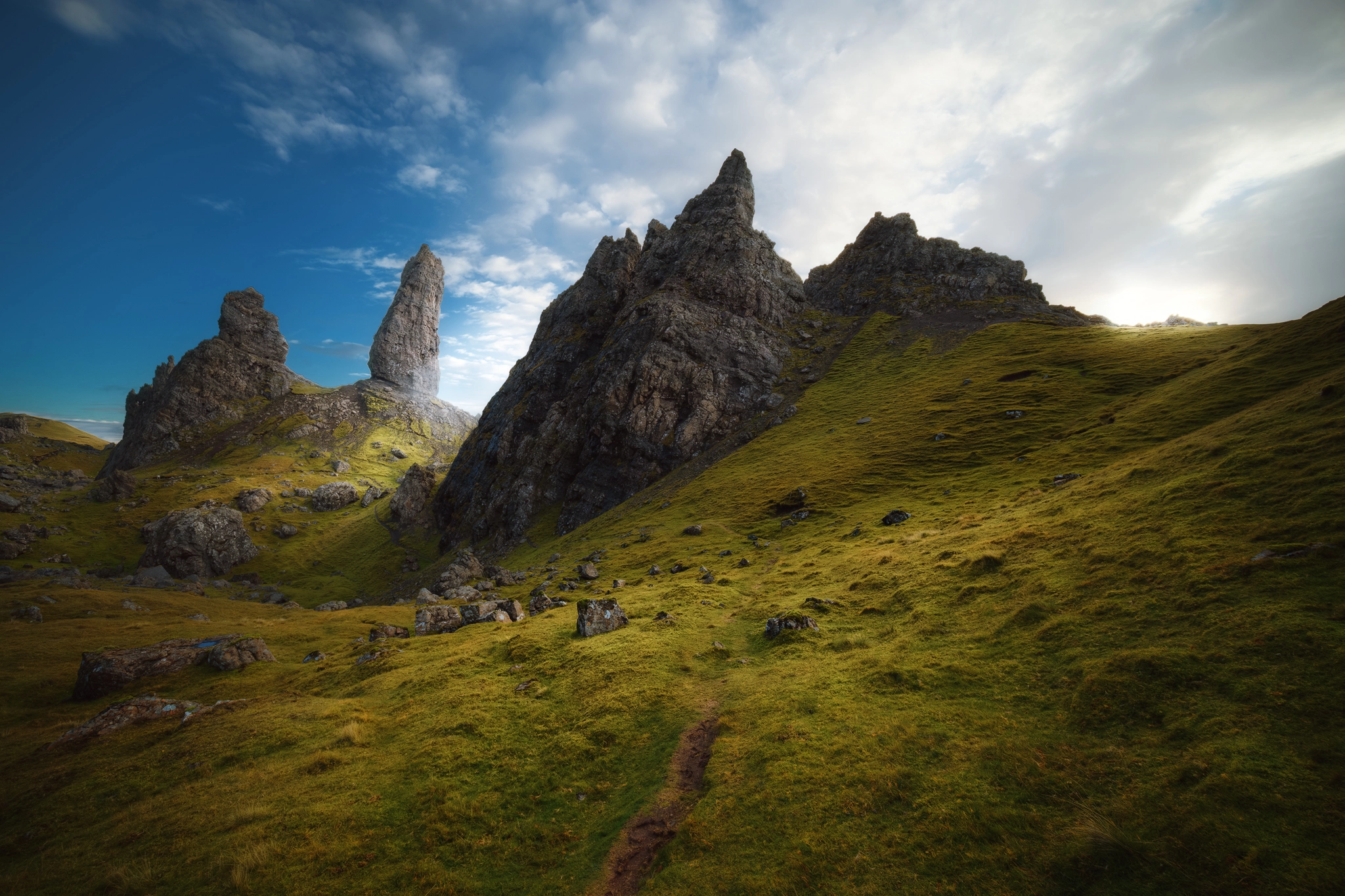 The dramatic landscape of the Storr on the Isle of Skye, Scotland, featuring towering, jagged rock formations rising sharply from a gently sloping hillside covered in vibrant green grass. A narrow, muddy path winds through the foreground, leading the viewer’s eye towards the iconic Old Man of Storr pinnacle, which is strikingly illuminated by soft sunlight breaking through a partially cloudy sky. Scattered rocks and boulders are strewn across the grassy slopes, enhancing the wild and ancient atmosphere of the scene.