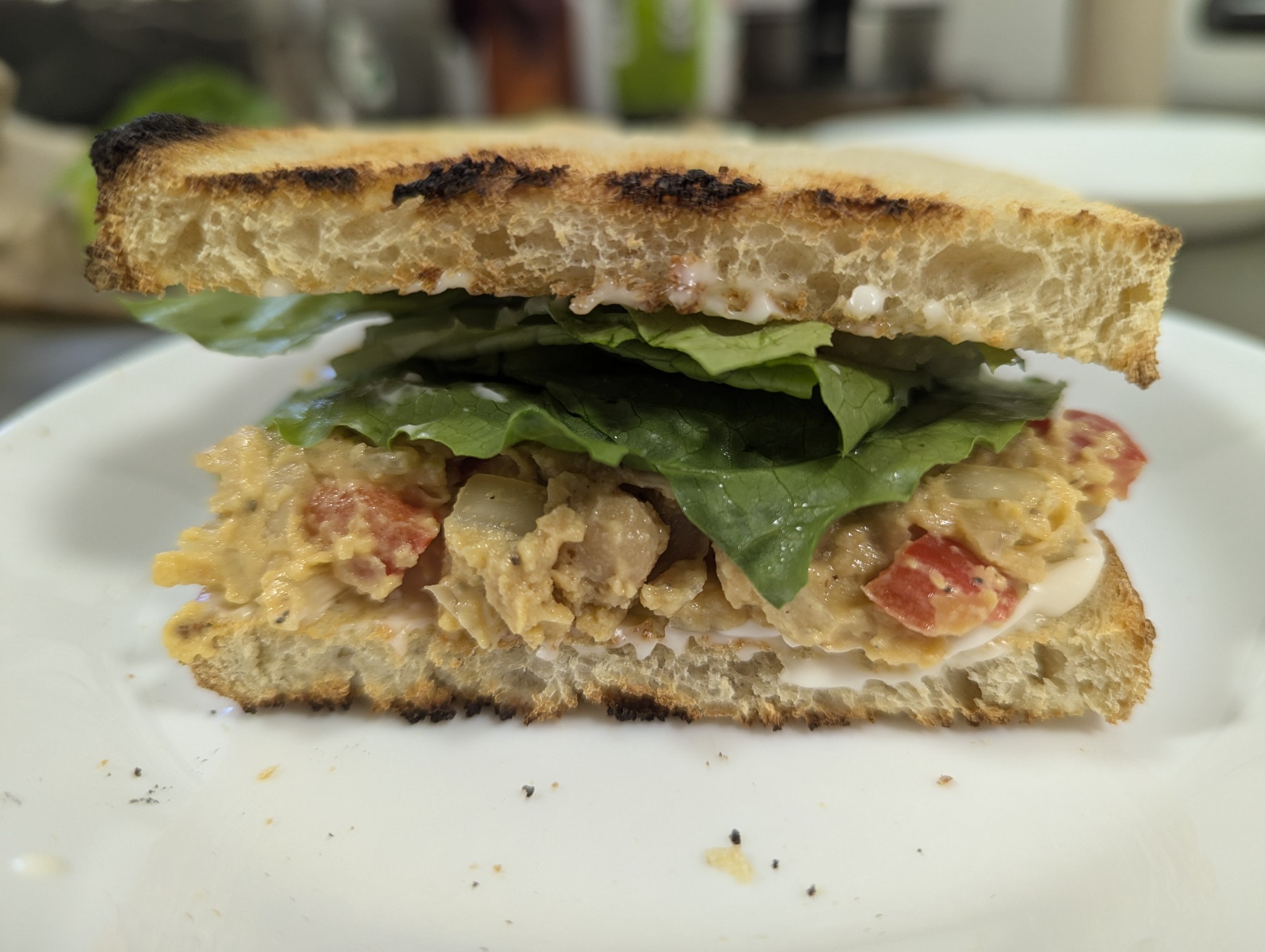 image contains a sandwich with toasted bread, lettuce, and a chickpea salad filling on a white plate and blurred background.
