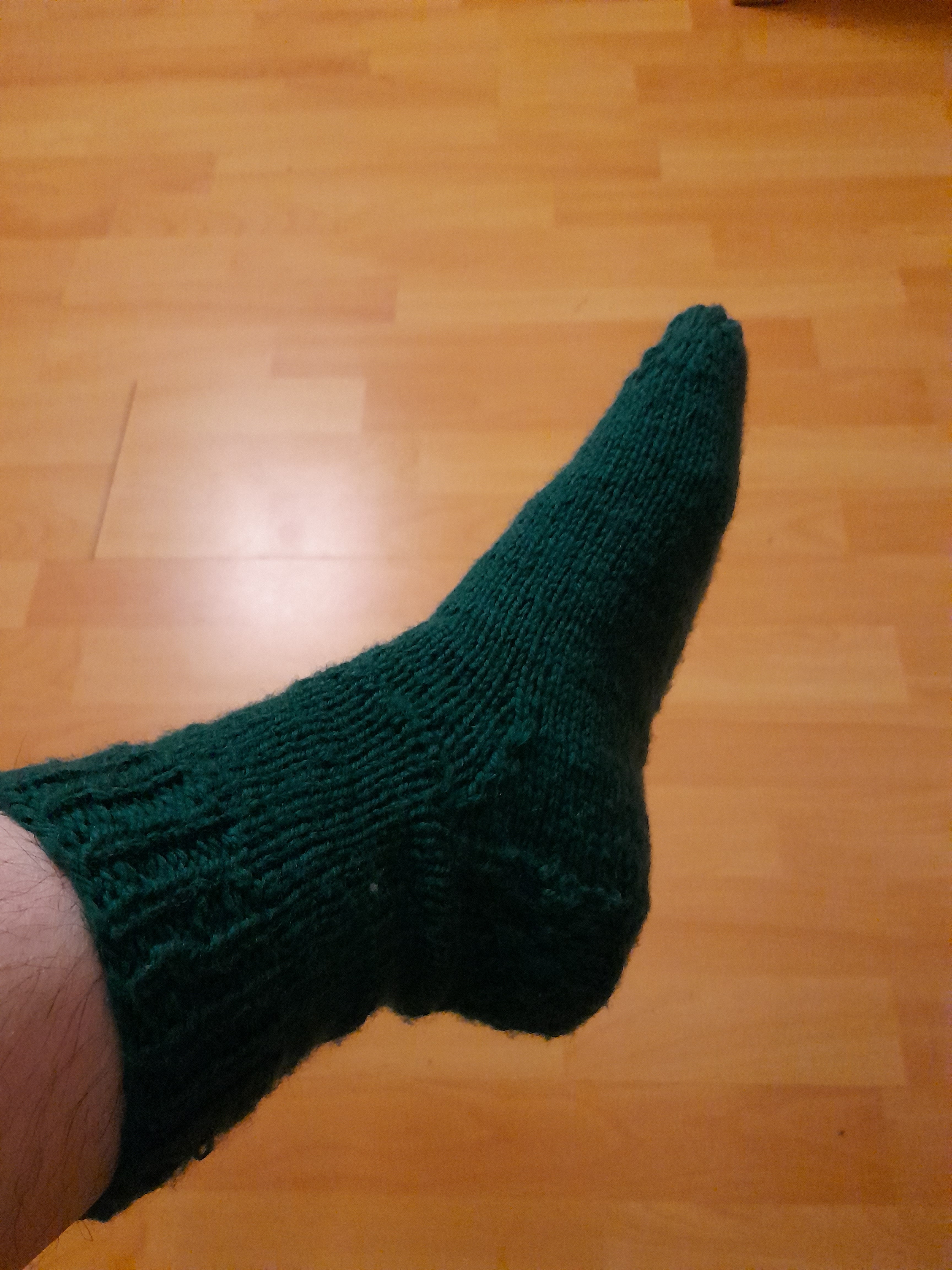 une chaussette verte tricotée main, la lumière est très jaune et on voit un bout de parquet clair derrière.