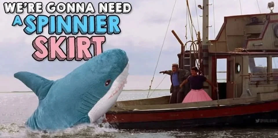 A picture from jaws showing blåhaj attacking the boat the Orca. Quint and Brody are aboard the ship with Brody wearing a pink skirt. The picture is captioned "We're gonna need a SPINNIER SKIRT"