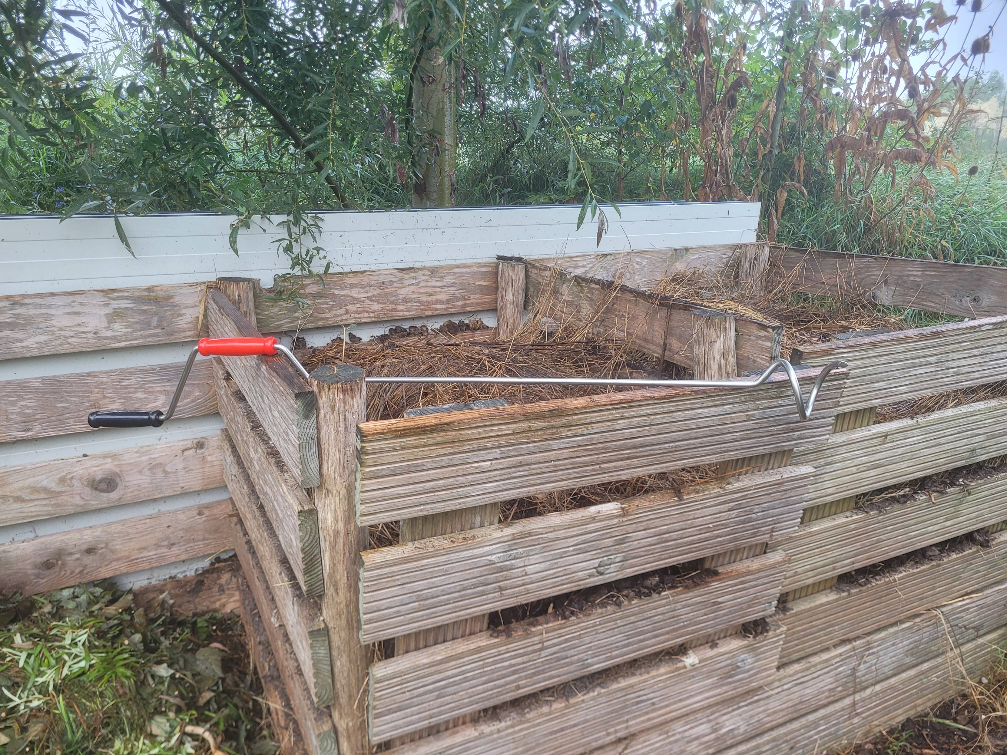 Houten compostbakken met daarop liggend een compostswoeler met een handvat als van een zonneluifel met een grijs en rood handvat