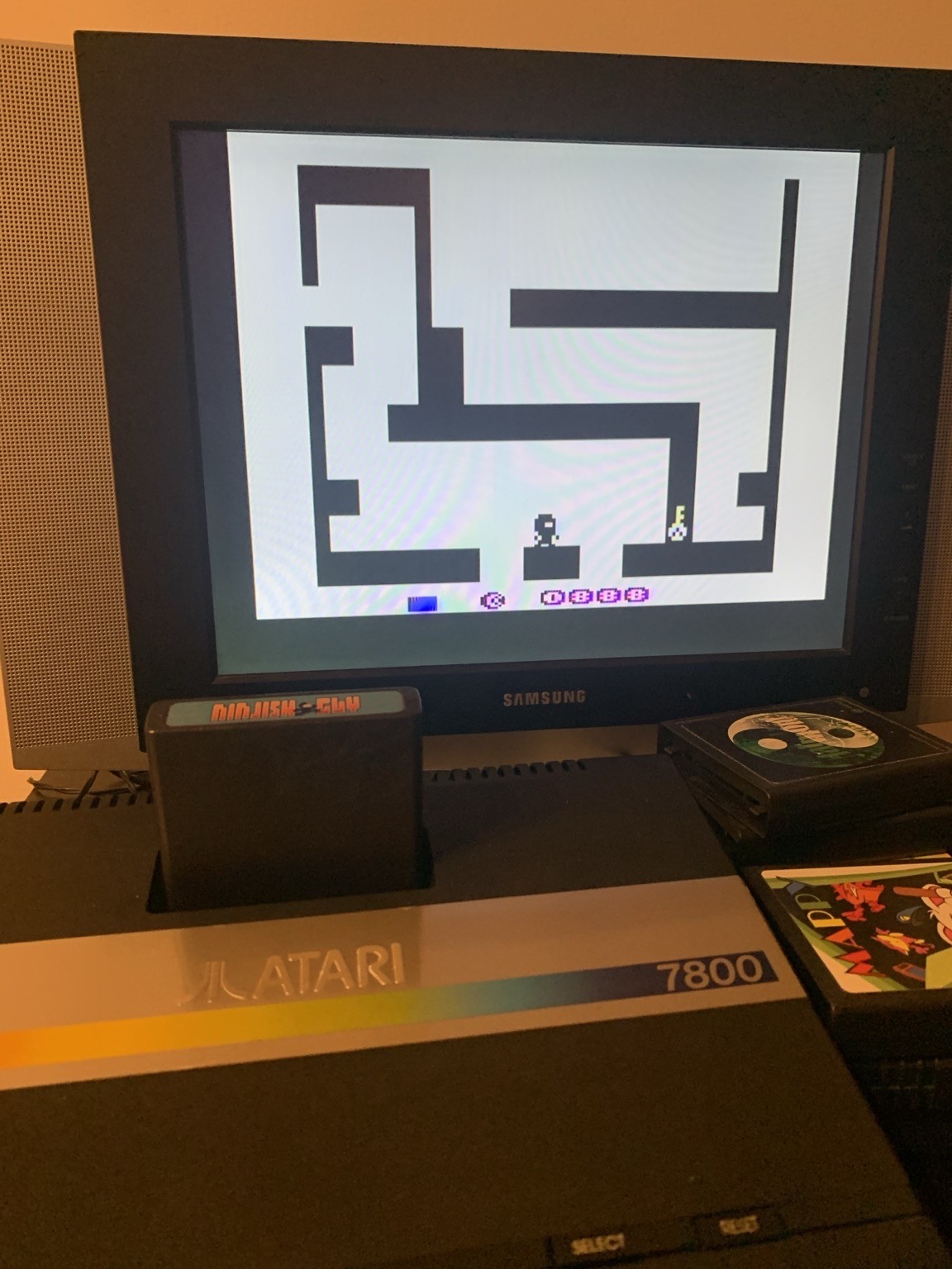 An Atari 7800 console displaying a black-and-white video game on a screen. The game features a character navigating a maze-like structure. An Atari game cartridge is inserted into the console, with additional game cases visible nearby.